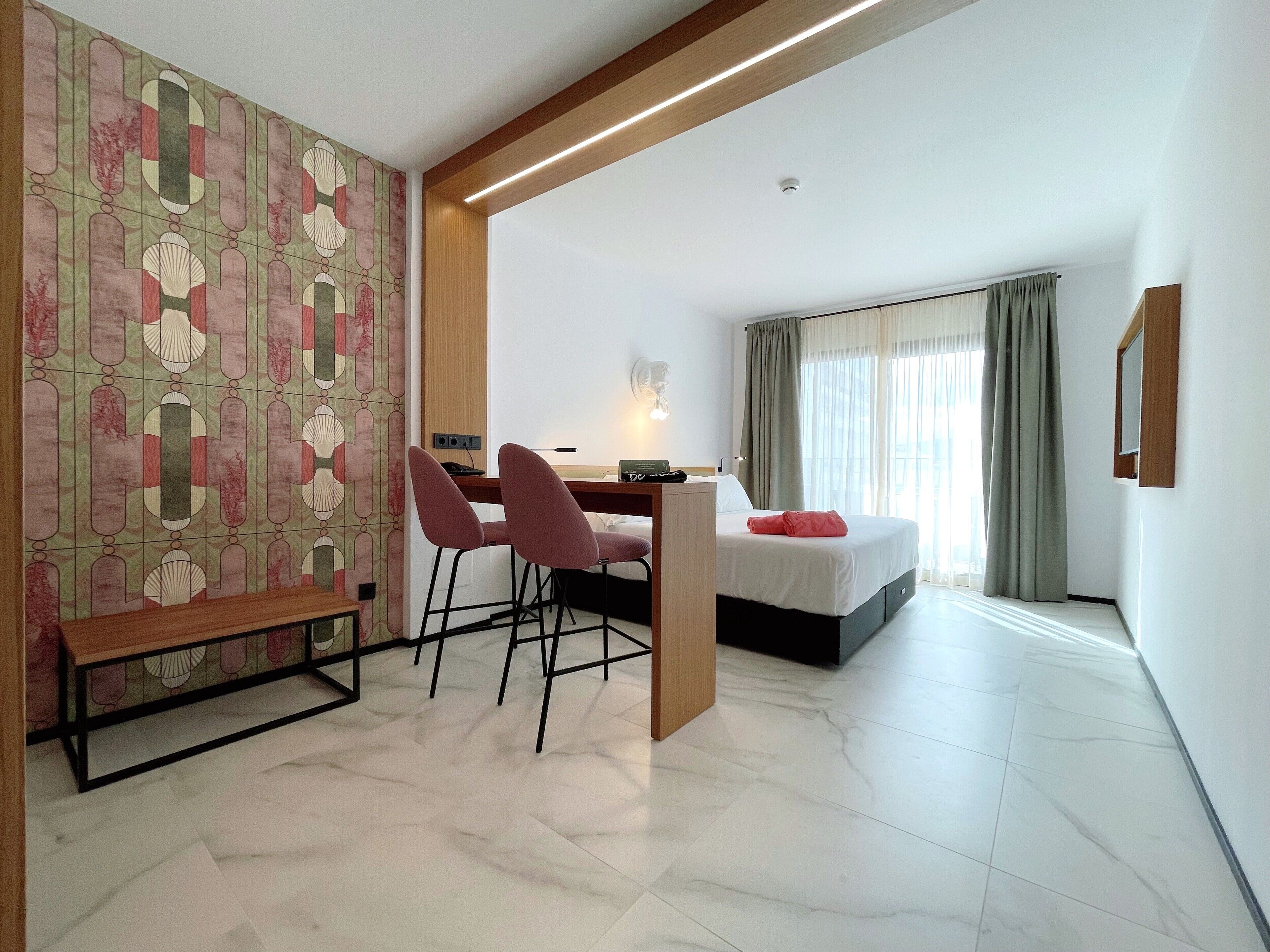 A hotel room with a bed, a desk with two chairs, a patterned wall, and a window with curtains.