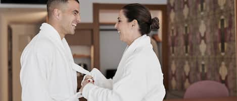 Two people in white bathrobes standing in a room with a wooden desk and a patterned wall.