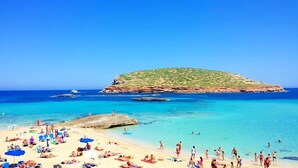 Una playa con gente tomando el sol y nadando, un afloramiento rocoso y un cielo azul despejado.