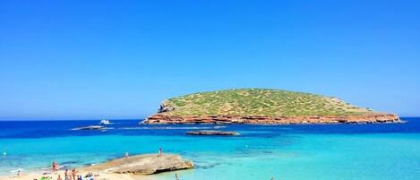 Una playa con gente tomando el sol y nadando, un afloramiento rocoso y un cielo azul despejado.