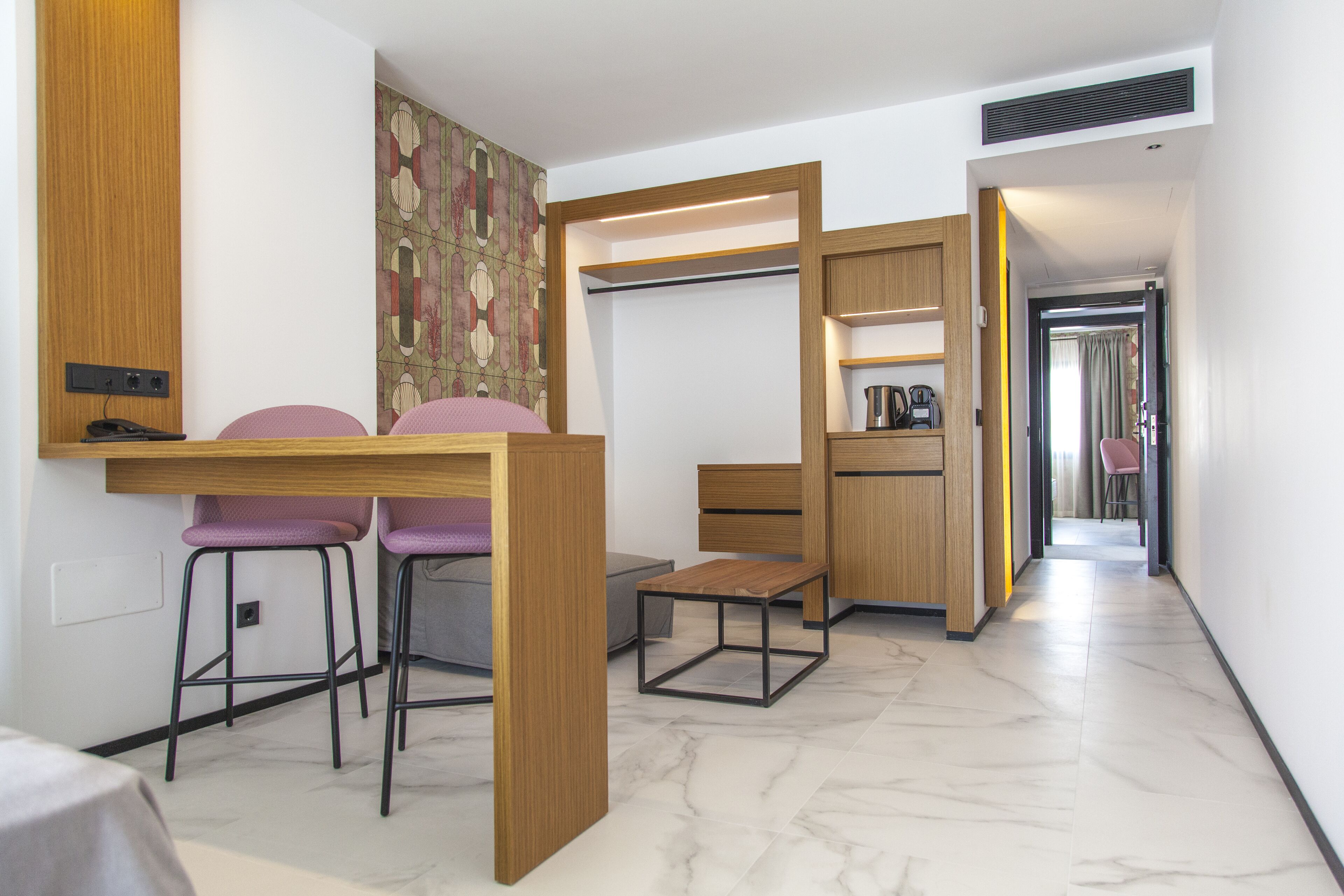A compact hotel room with a wooden desk, two purple chairs, a small table, and a kitchenette area.