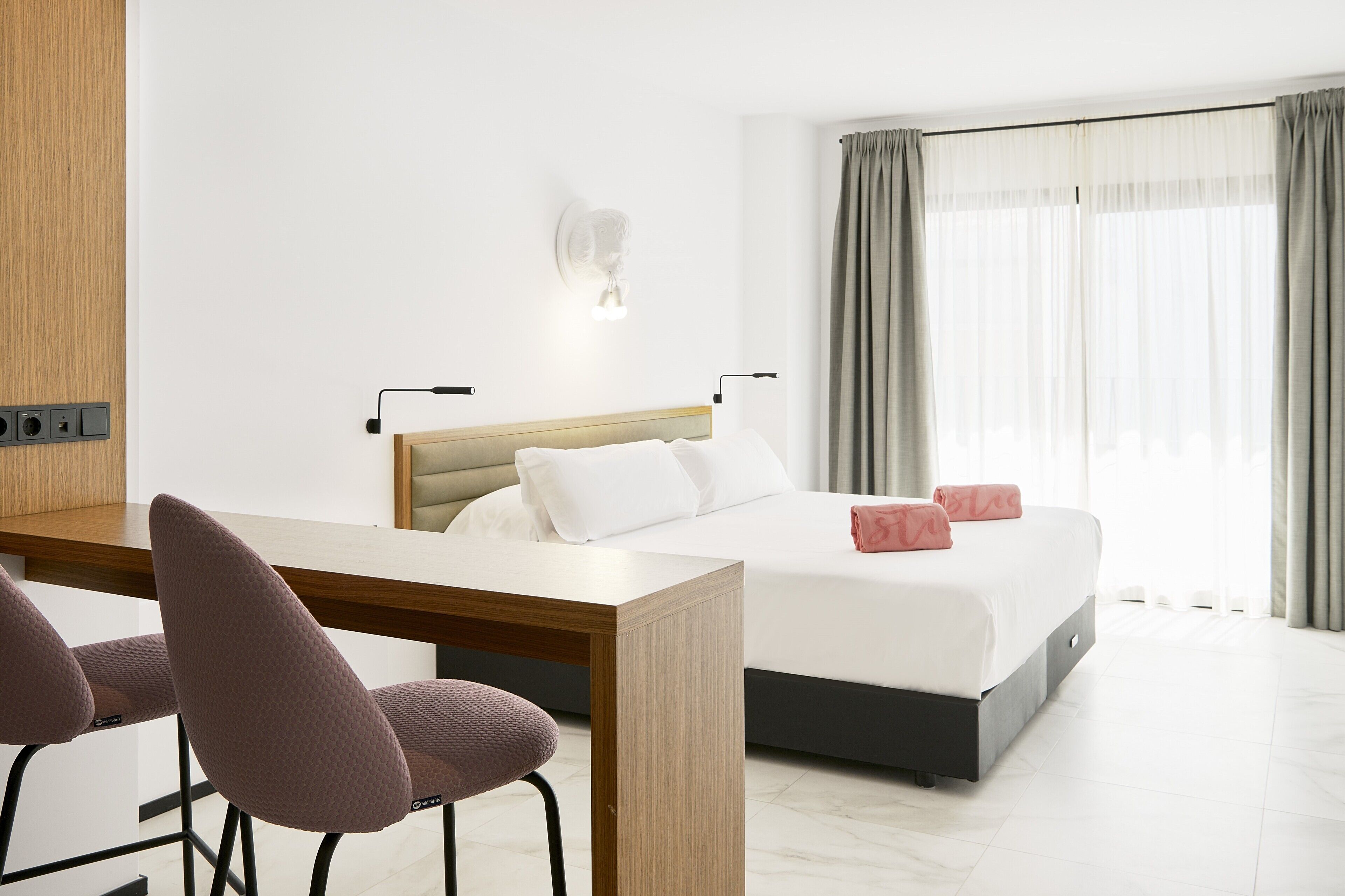 A hotel room with a bed, a desk, two chairs, a wall-mounted light, and a window with curtains.