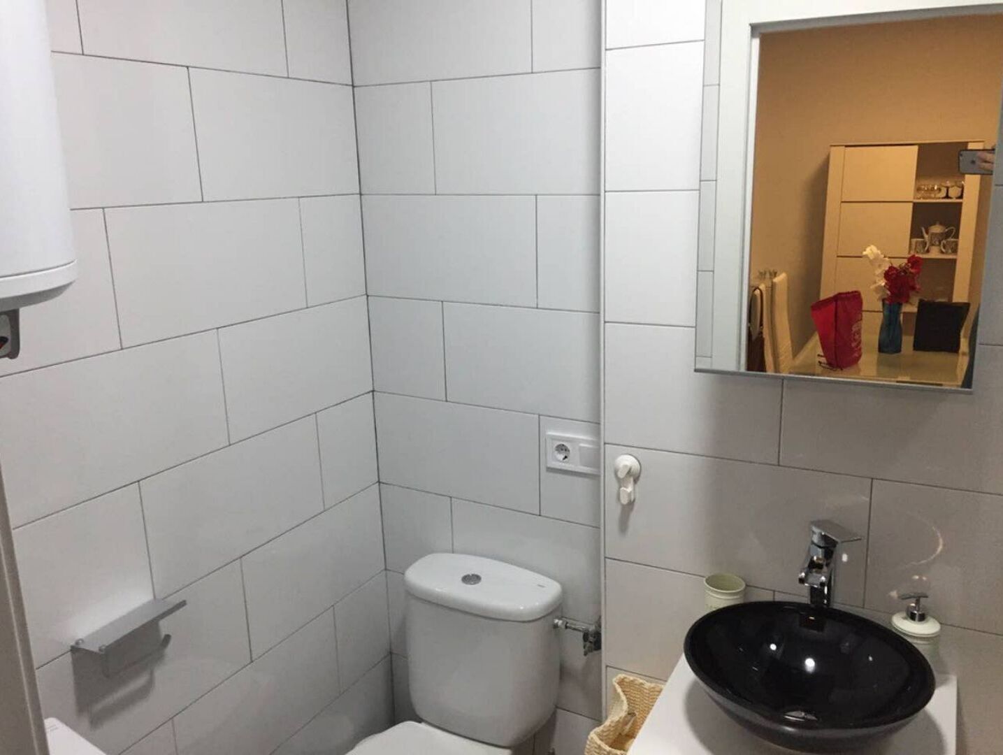 Un baño pequeño y moderno con azulejos blancos, un inodoro, un lavabo con un lavamanos negro y un espejo sobre el lavabo.