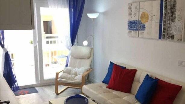 A modern living room with a white sofa, a blue coffee table, and a white armchair. There is a painting on the wall and a window with blue curtains. - Downtown beach & peace (Málaga)