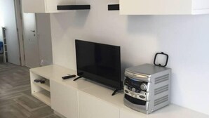 A modern living room with a flat-screen TV on a white entertainment unit, a small table with a blue object, and a grey speaker. - Downtown beach & peace (Málaga)