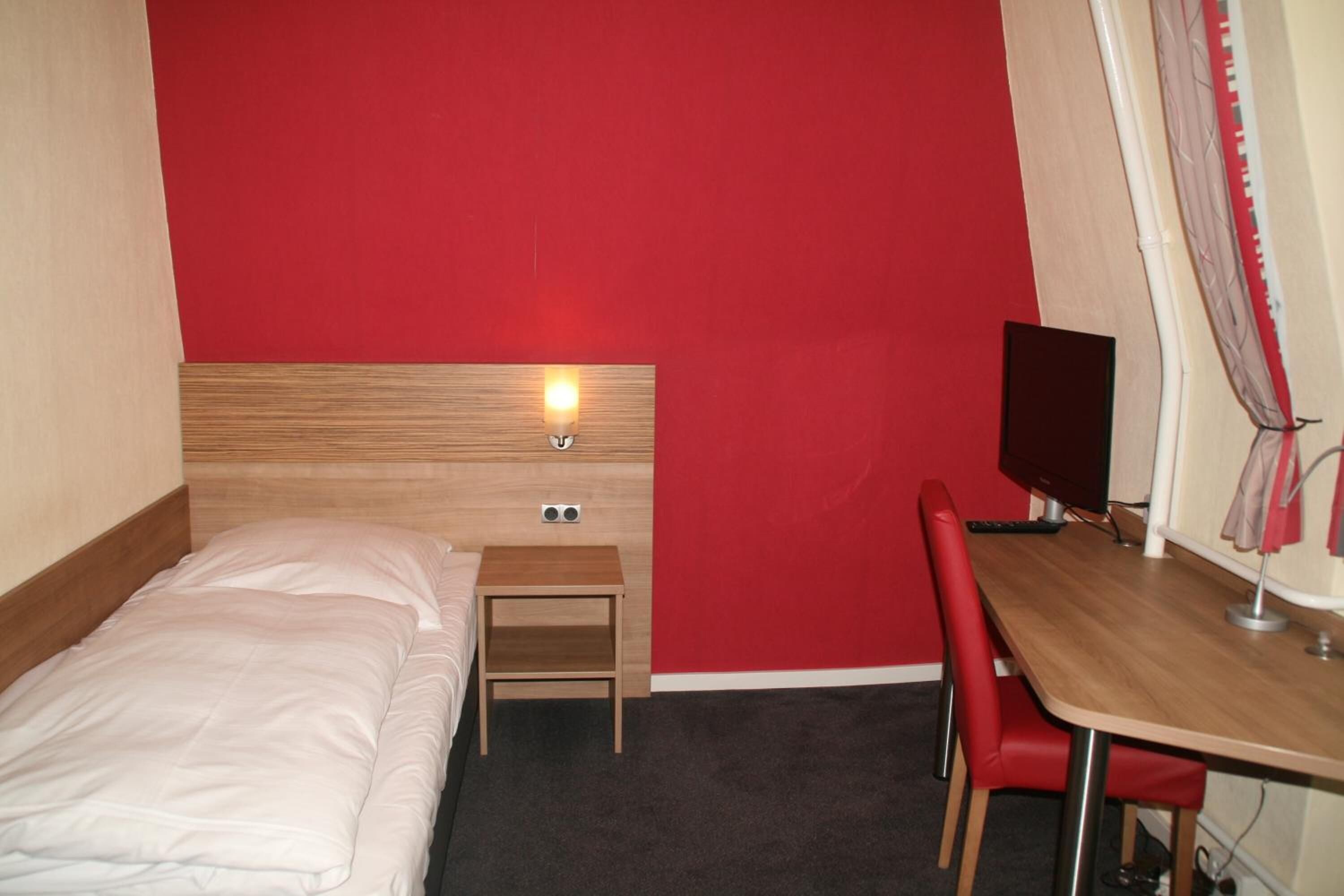 Quarto de hotel com cama, escrivaninha, cadeira, televisão e parede vermelha.