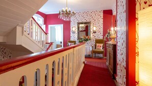Una escalera con barandilla roja que conduce a un piso superior. Un candelabro cuelga del techo y un espejo está fijado a la pared.