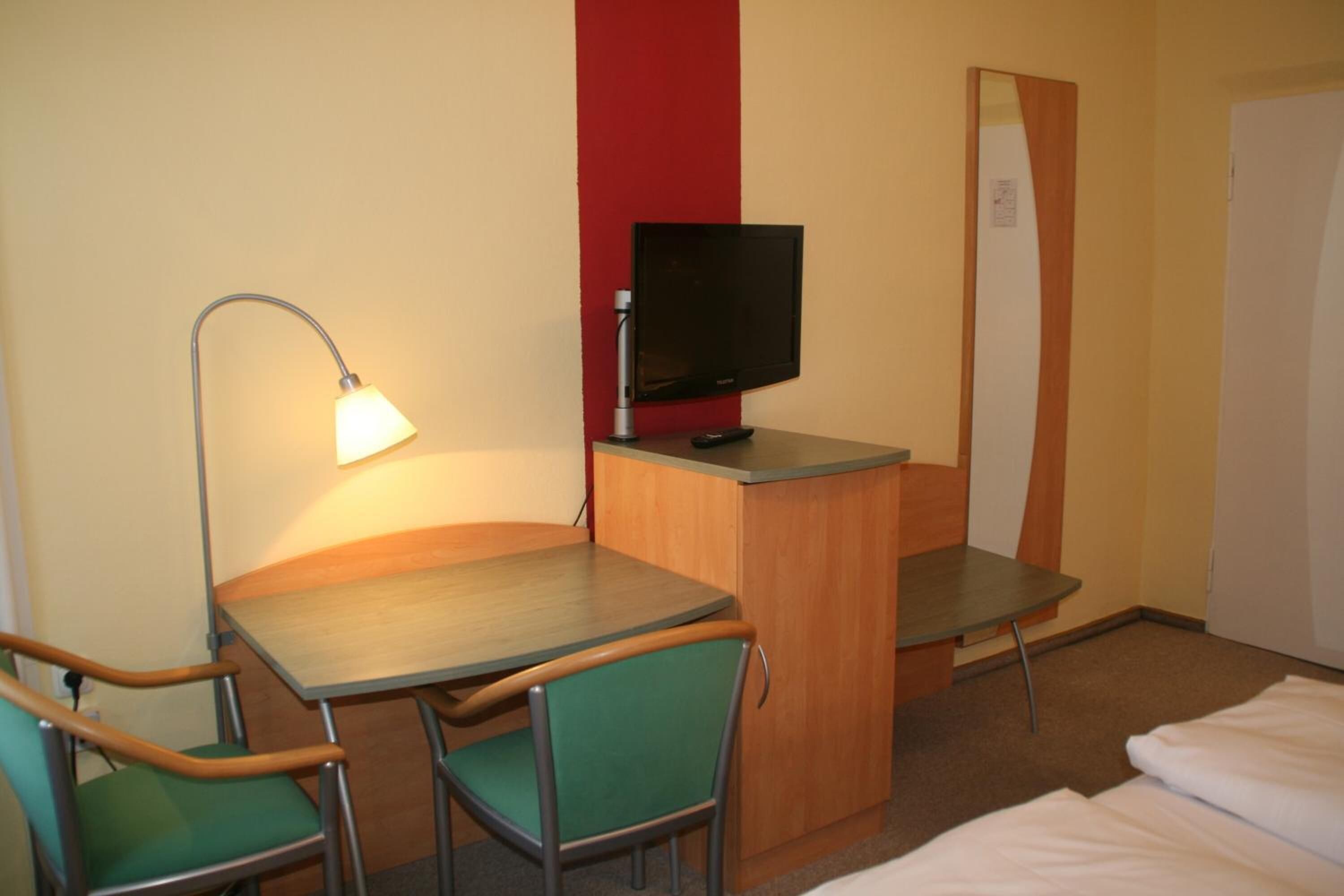 A hotel room with a desk, two chairs, a television, a lamp, and a bed.