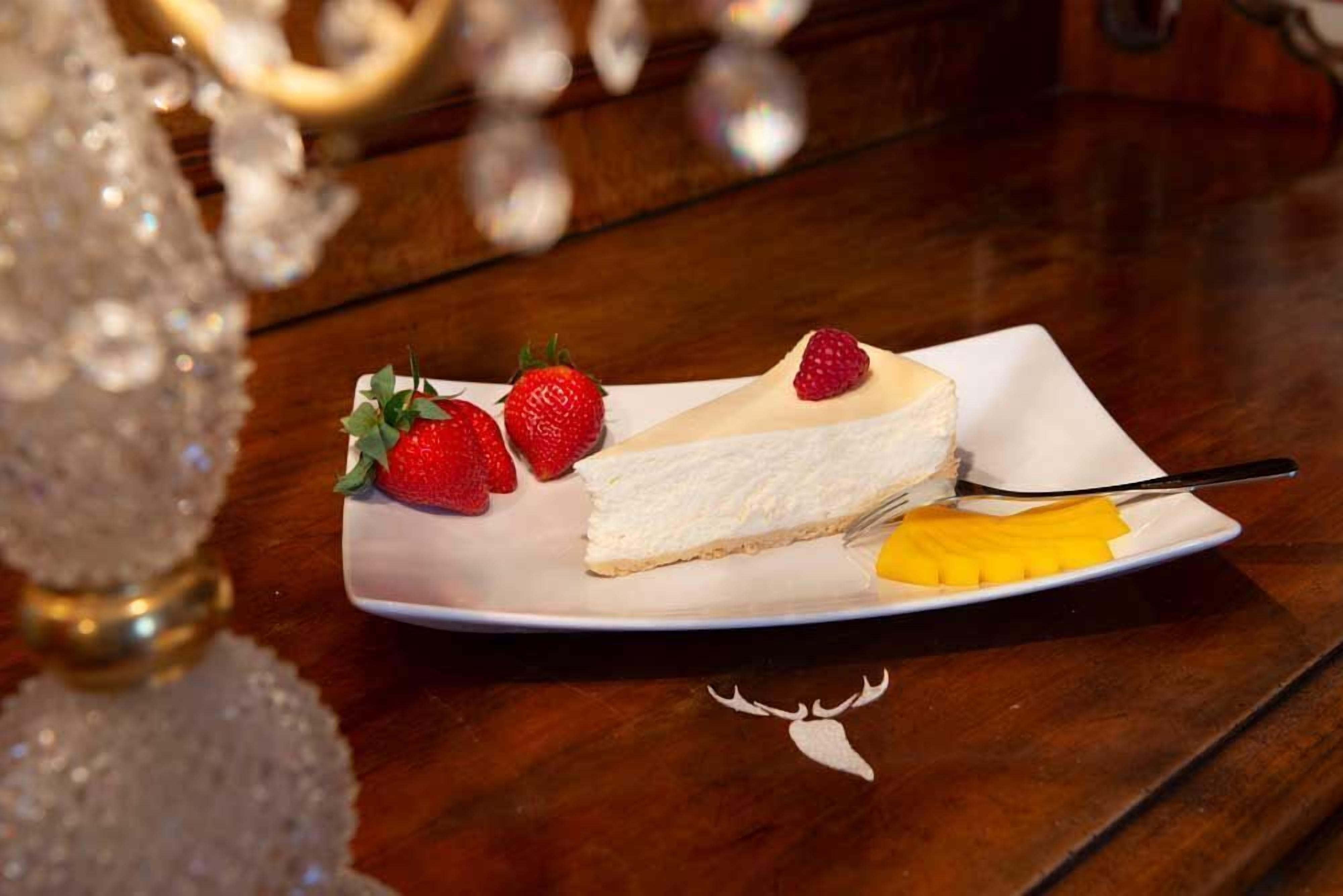 Una porció de pastís de formatge amb maduixes i mango sobre un plat blanc, damunt d'una taula de fusta.