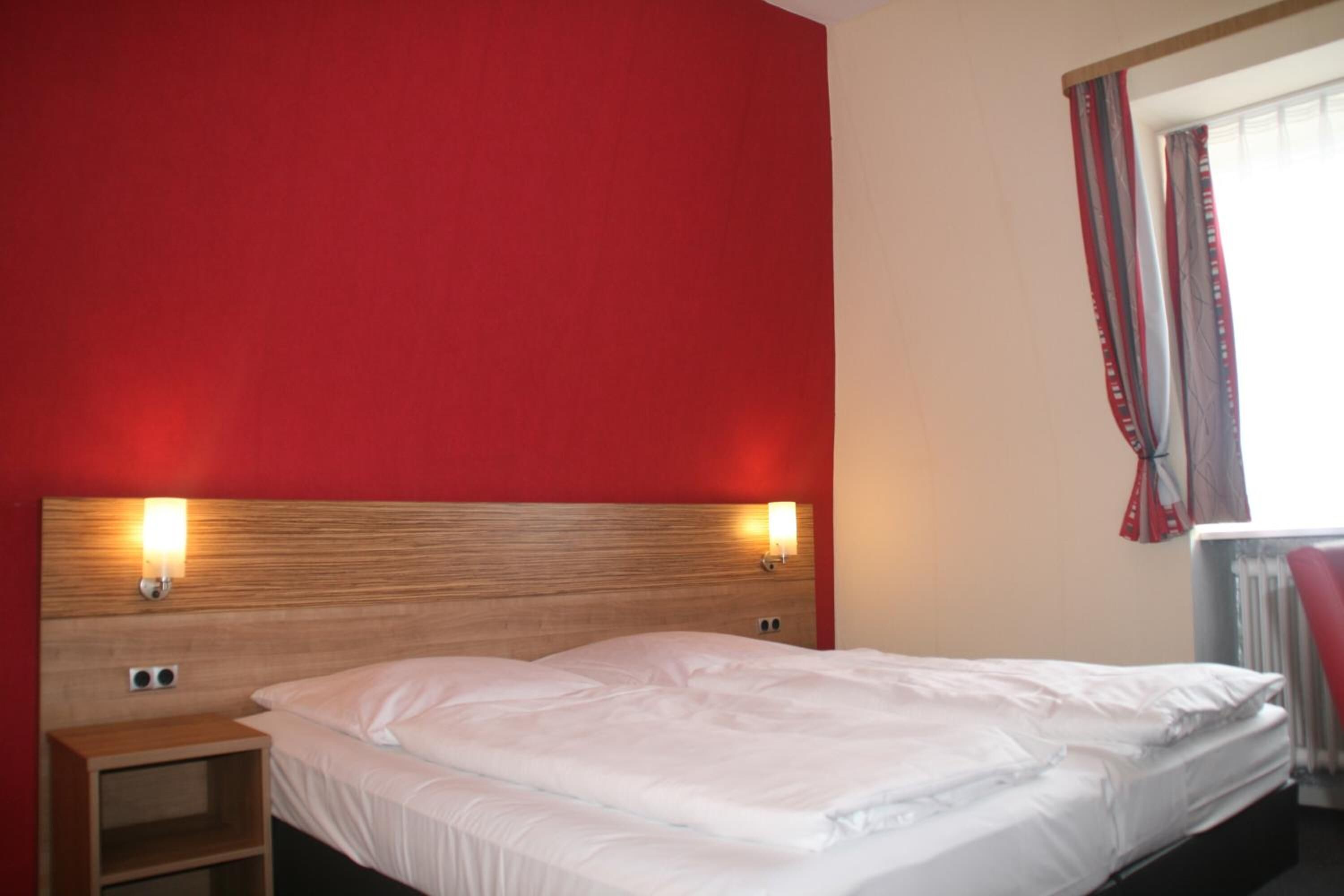 Hotelkamer met houten hoofdeinde, twee bedlampen, een bed met witte lakens en een raam met rode gordijnen.
