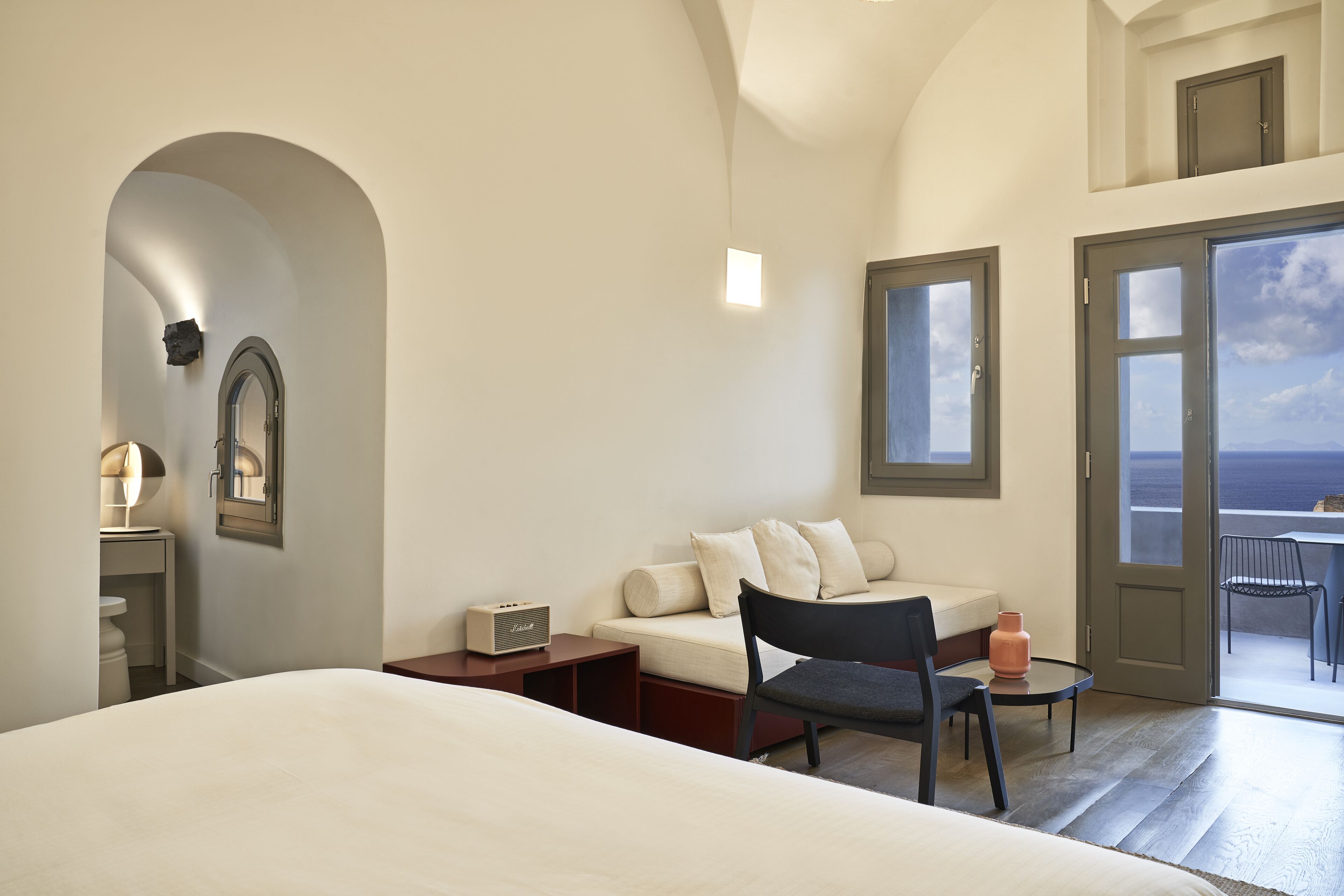 A modern hotel room with a bed, a sofa, a chair, a small table, a radio, and a view of the sea through the door.