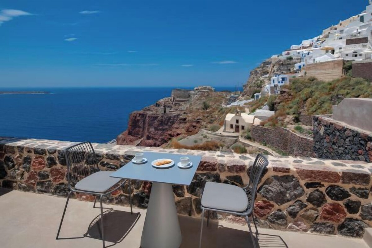 Un espace extérieur aménagé avec des murs en pierre, comprenant une table et des chaises, et offrant une vue sur un paysage côtier avec des bâtiments blancs et la mer.