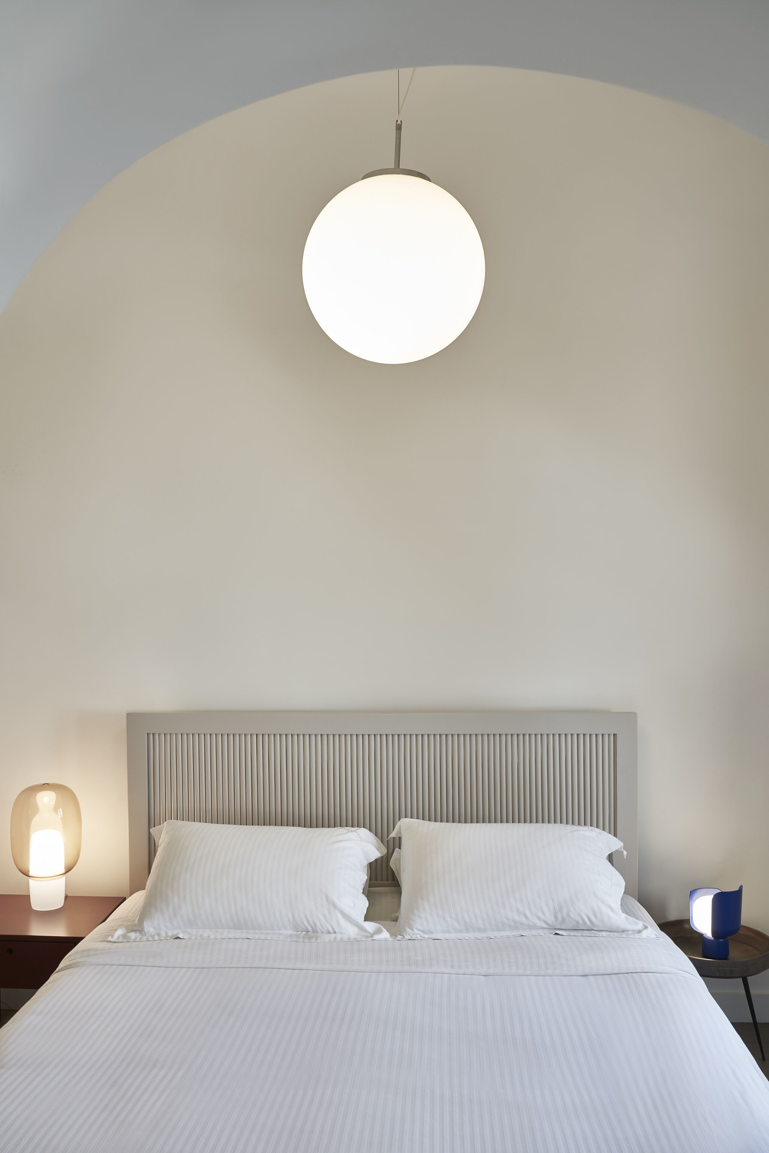 A neatly made bed with white linens, a round ceiling light, and a bedside table with a lamp.