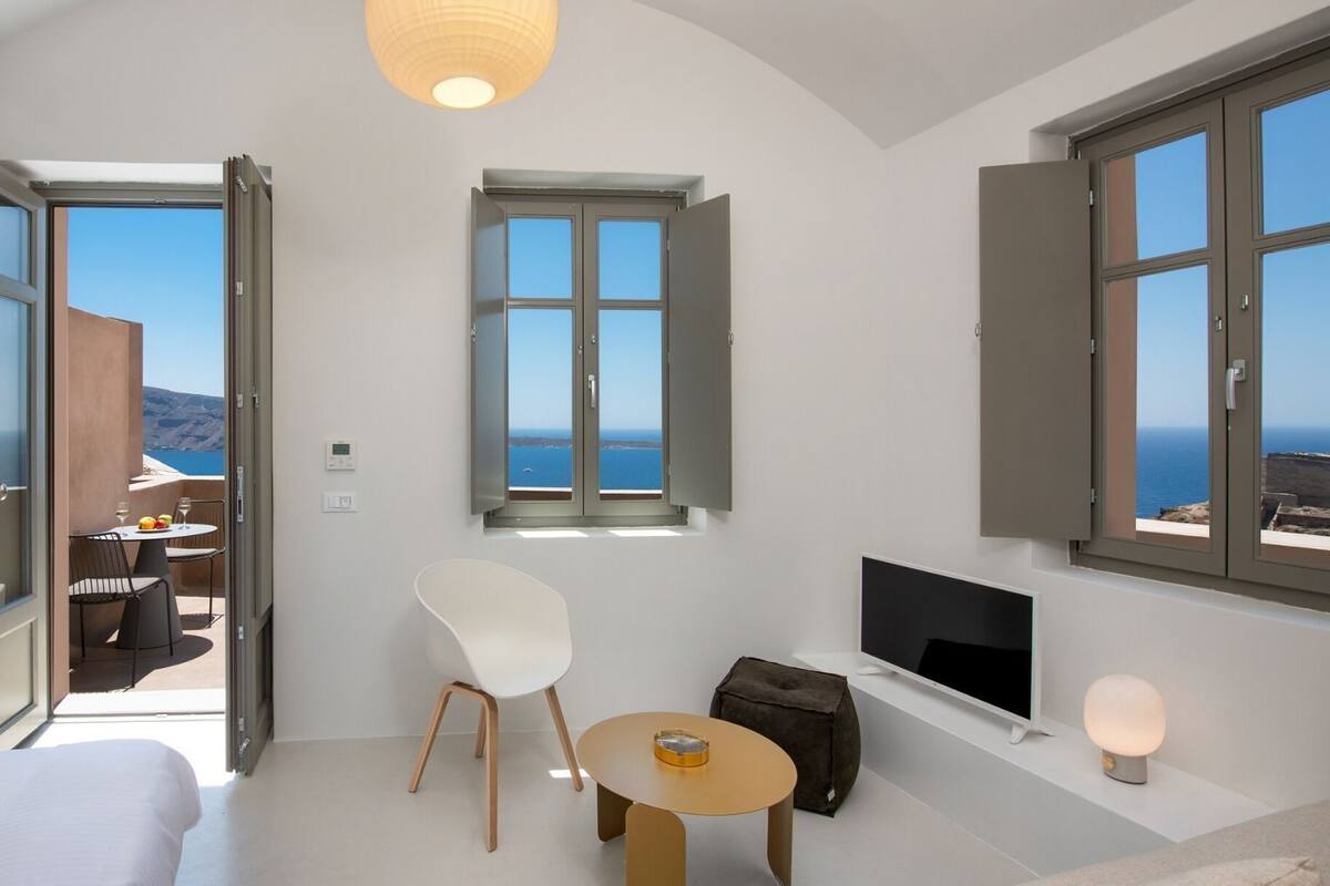 A modern hotel room with a bed, a TV, a small table, and a chair. There are two windows offering a view of the sea.