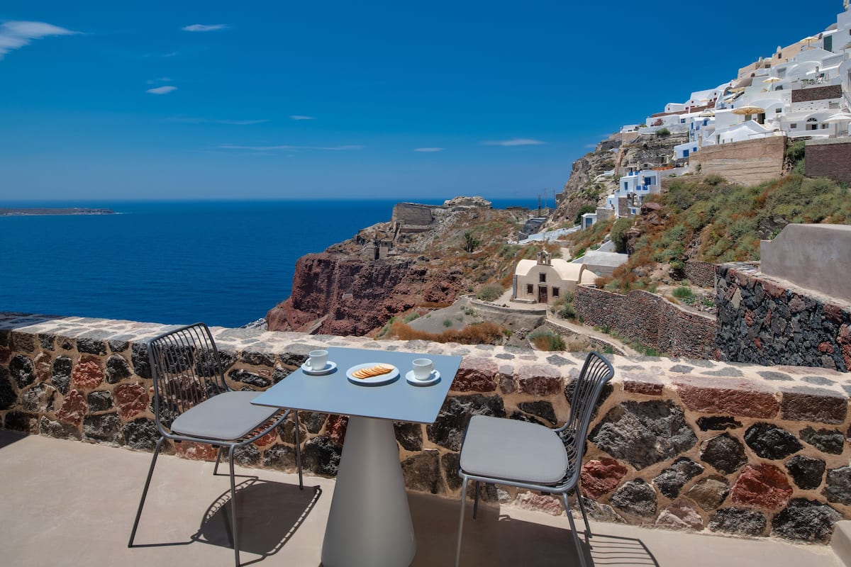 A table with two chairs and a plate of food on a terrace overlooking a coastal landscape with white buildings and a view of the sea.