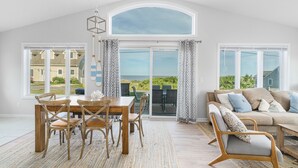 A spacious living room with a high ceiling, large windows, a dining area with wooden furniture, and a view of the outdoors. - 6 Bedroom House, Ocean View, A/C, One Floor, Less than 1 Mile to Beach (Narragansett)