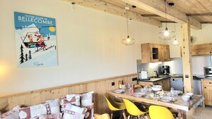 A cozy dining area with a wooden table set for four, yellow chairs, and a patterned sofa. A kitchen with wooden cabinets and modern appliances is visible in the background. - Quiet apartment near slopes, 6 ppl, parking (Notre-Dame-de-Bellecombe)