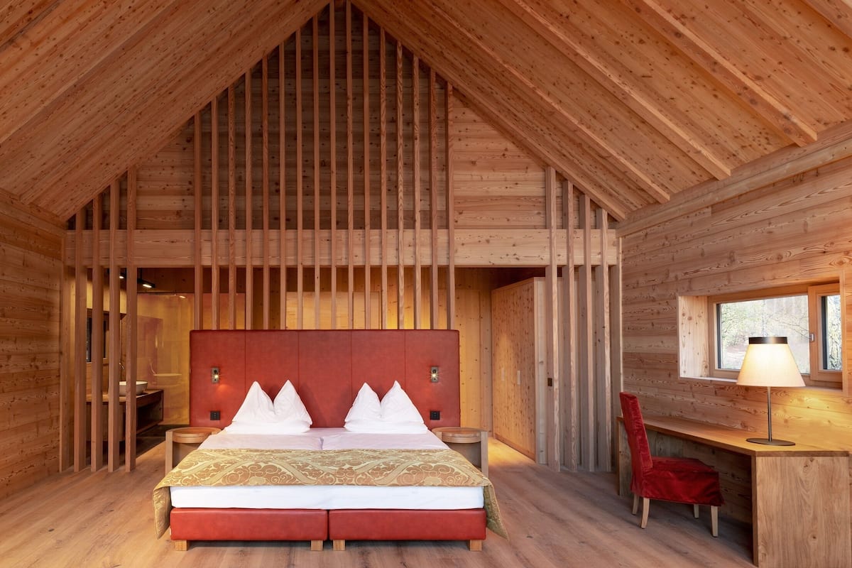 Une chambre dans une cabane en bois, avec un plafond haut, un lit doté d’une tête de lit rouge, un bureau, une chaise et une fenêtre.