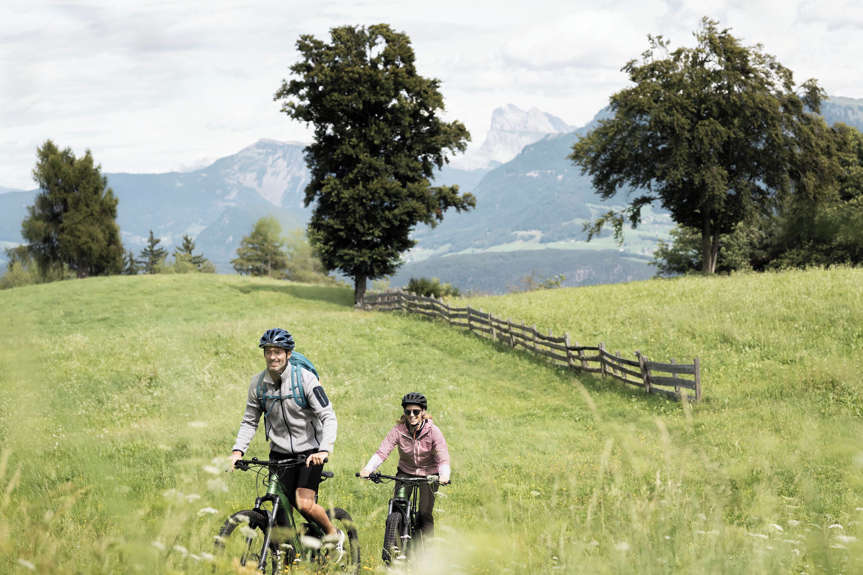 Two people on bicycles in a grassy field with a wooden fence and trees, and mountains in the background.