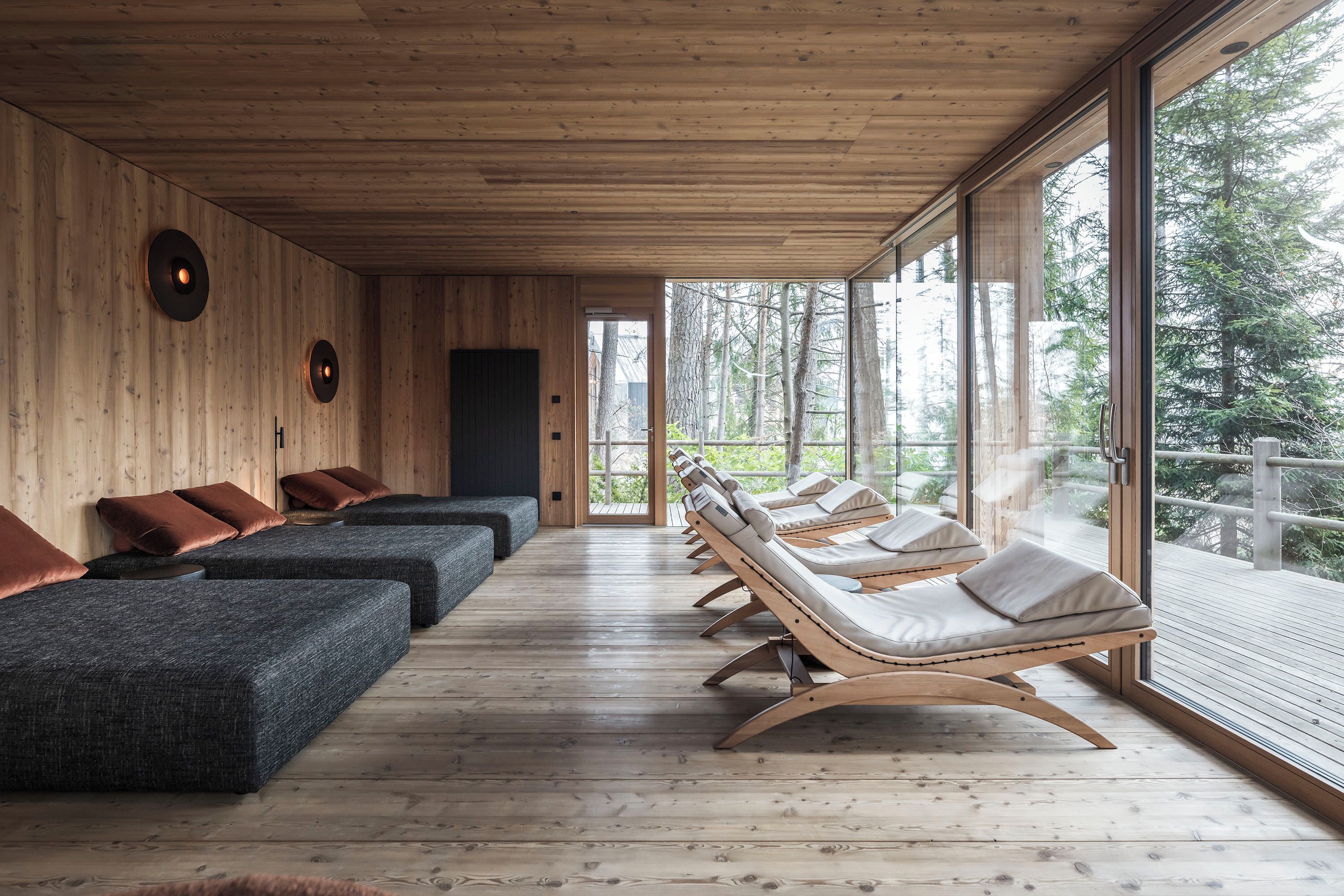 A row of lounge chairs with cushions, wooden walls, and a large window with a view of trees.