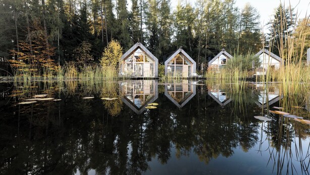 Three wooden cabins with glass windows, surrounded by tall trees and a calm lake reflecting the structures.