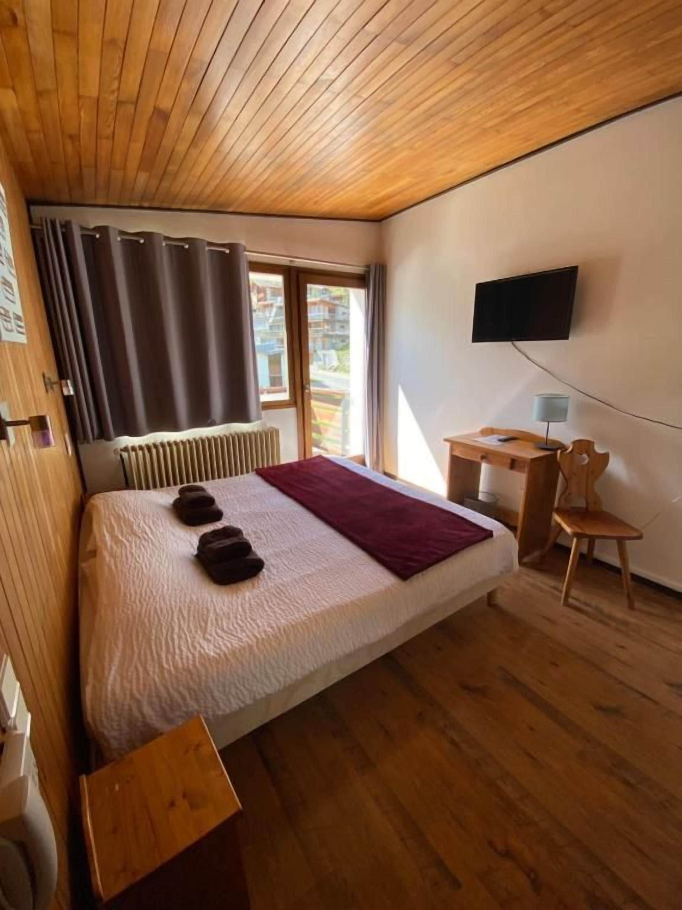 Un dormitorio con cama, una silla, un escritorio, un televisor y una ventana con cortinas.