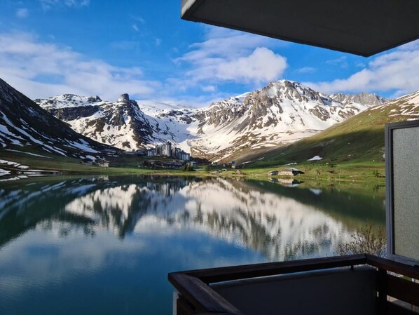 A mountain landscape with a lake reflecting the scenery, a clear sky, and buildings in the distance.