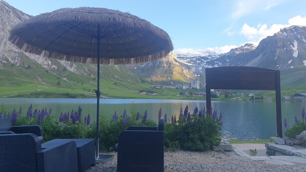 Une terrasse avec des sièges extérieurs, un parasol en chaume et une vue sur un lac, avec des montagnes en arrière-plan.