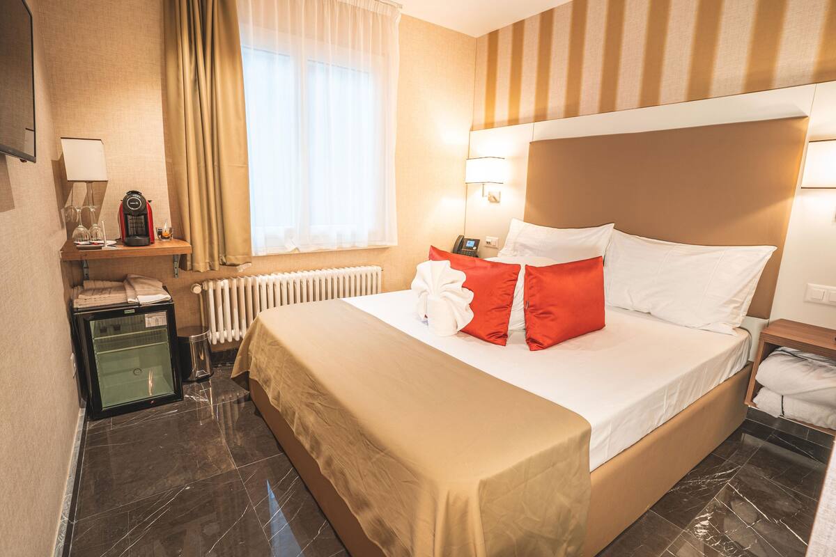 A hotel room with a large bed, a bedside table with a lamp, a coffee maker, and a view of the outside through a window with curtains.