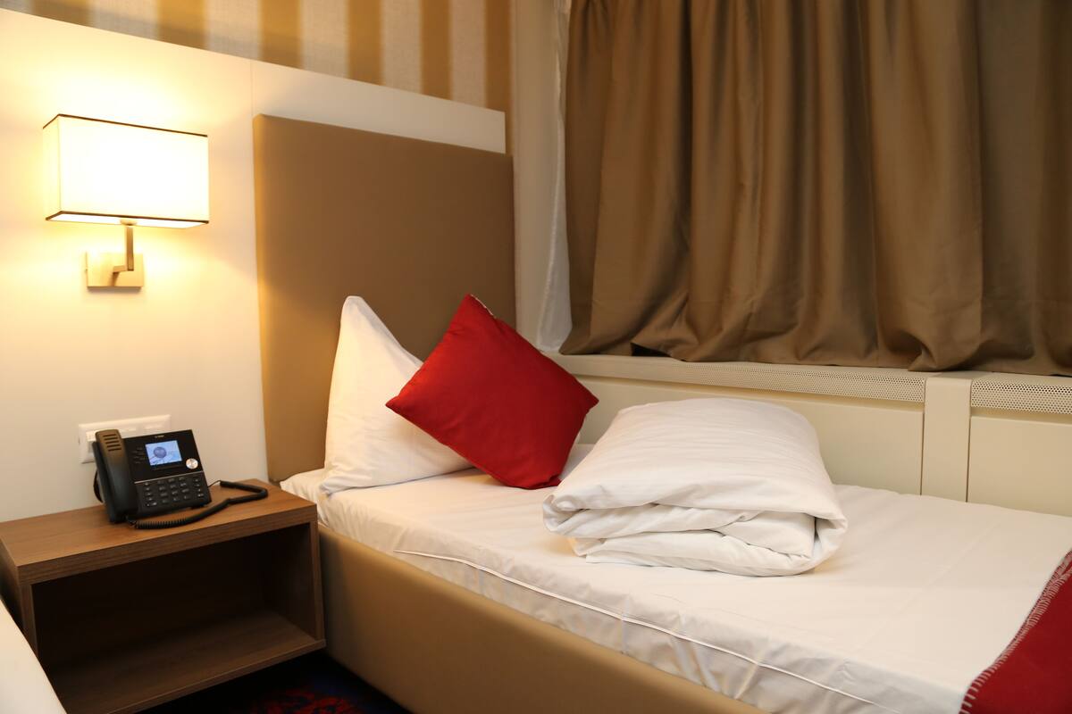 A hotel room with a bed, a nightstand with a phone, a window with curtains, and a wall-mounted lamp.