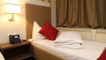 A hotel room with a bed, a nightstand with a phone, a window with curtains, and a wall-mounted lamp.