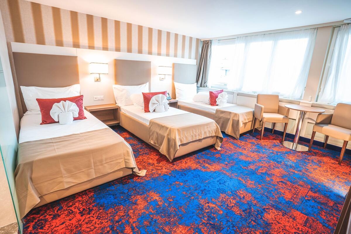 A hotel room with two beds, a small table, a chair, and a window with curtains.