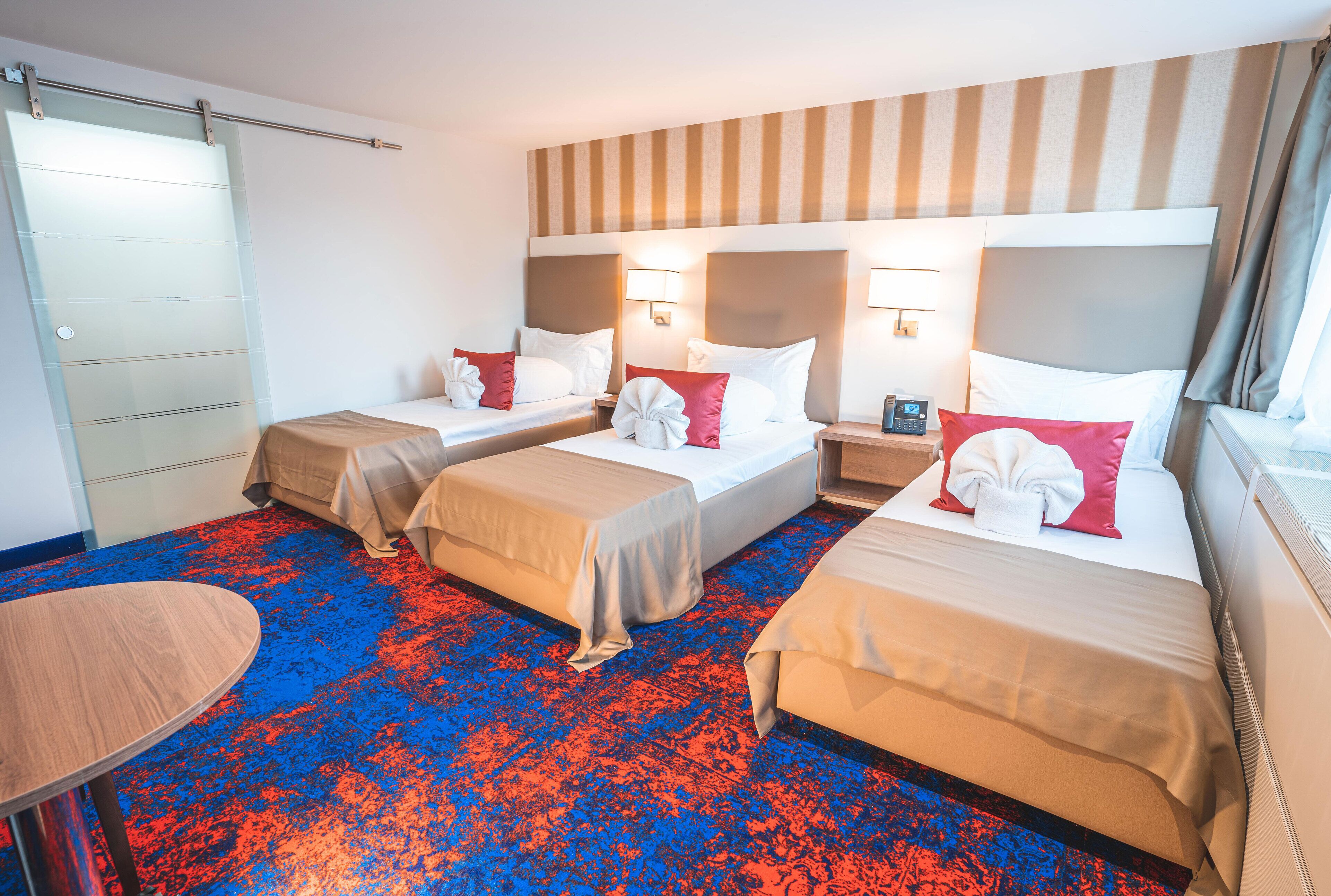 Una habitación de hotel con tres camas, una mesita pequeña y un ventanal grande.