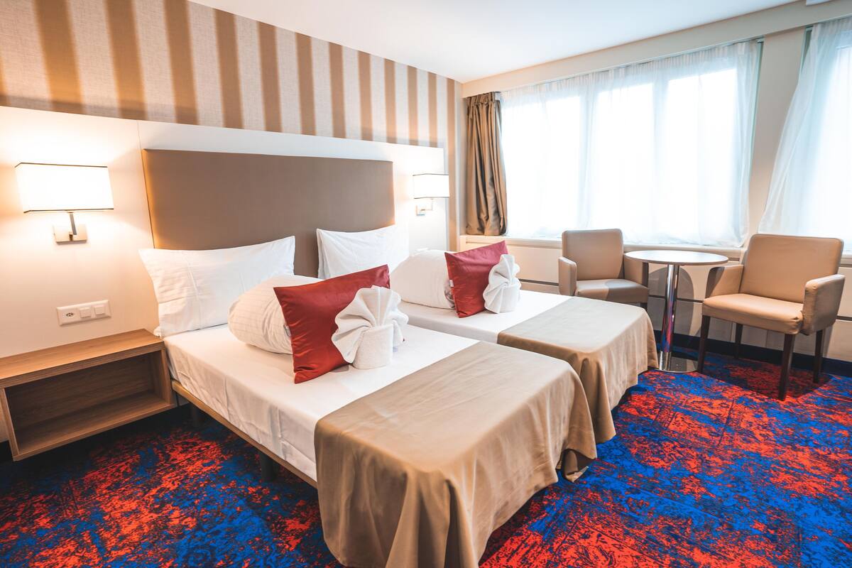 A hotel room with two beds, a small table, a chair, and a window with curtains.
