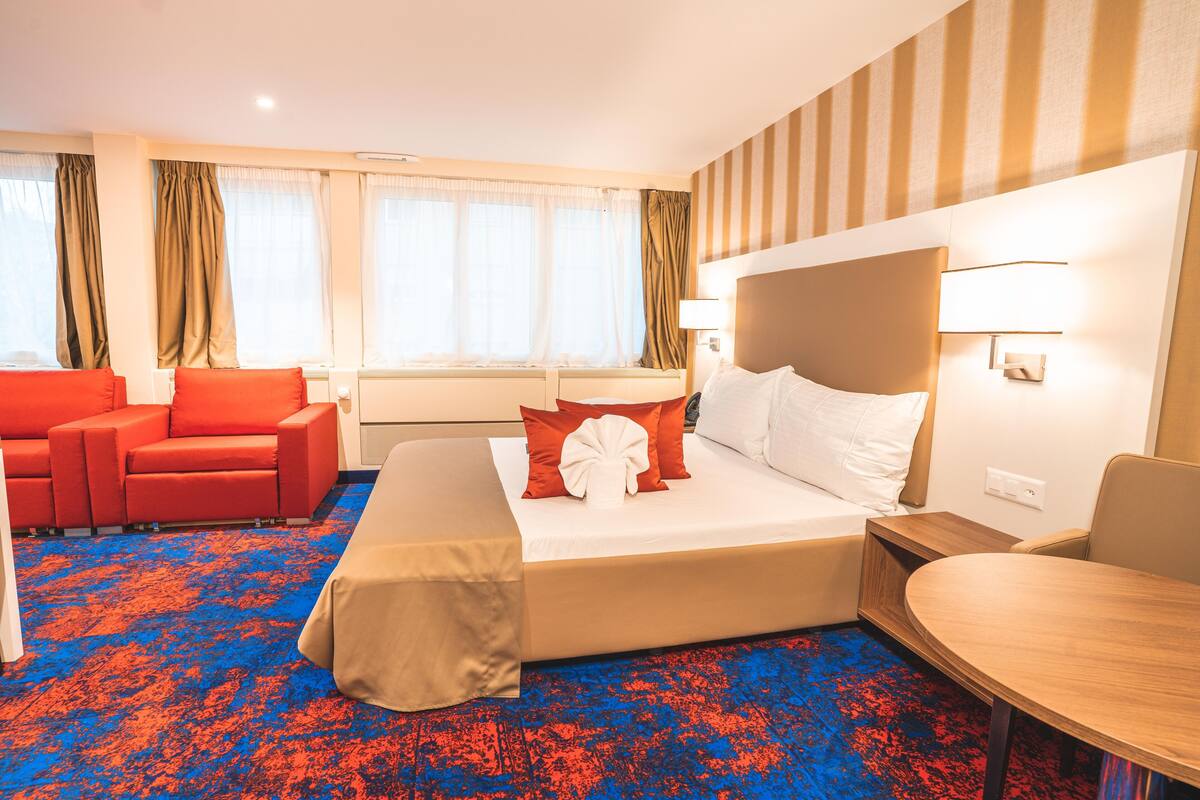 A hotel room with a large bed, a red sofa, a small table, and a window with curtains.