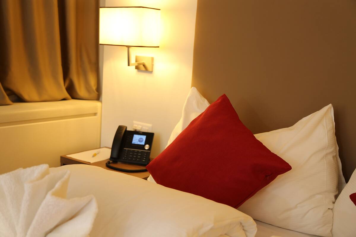 A hotel room with a bed, a bedside table with a phone, a lamp, and a window with curtains.