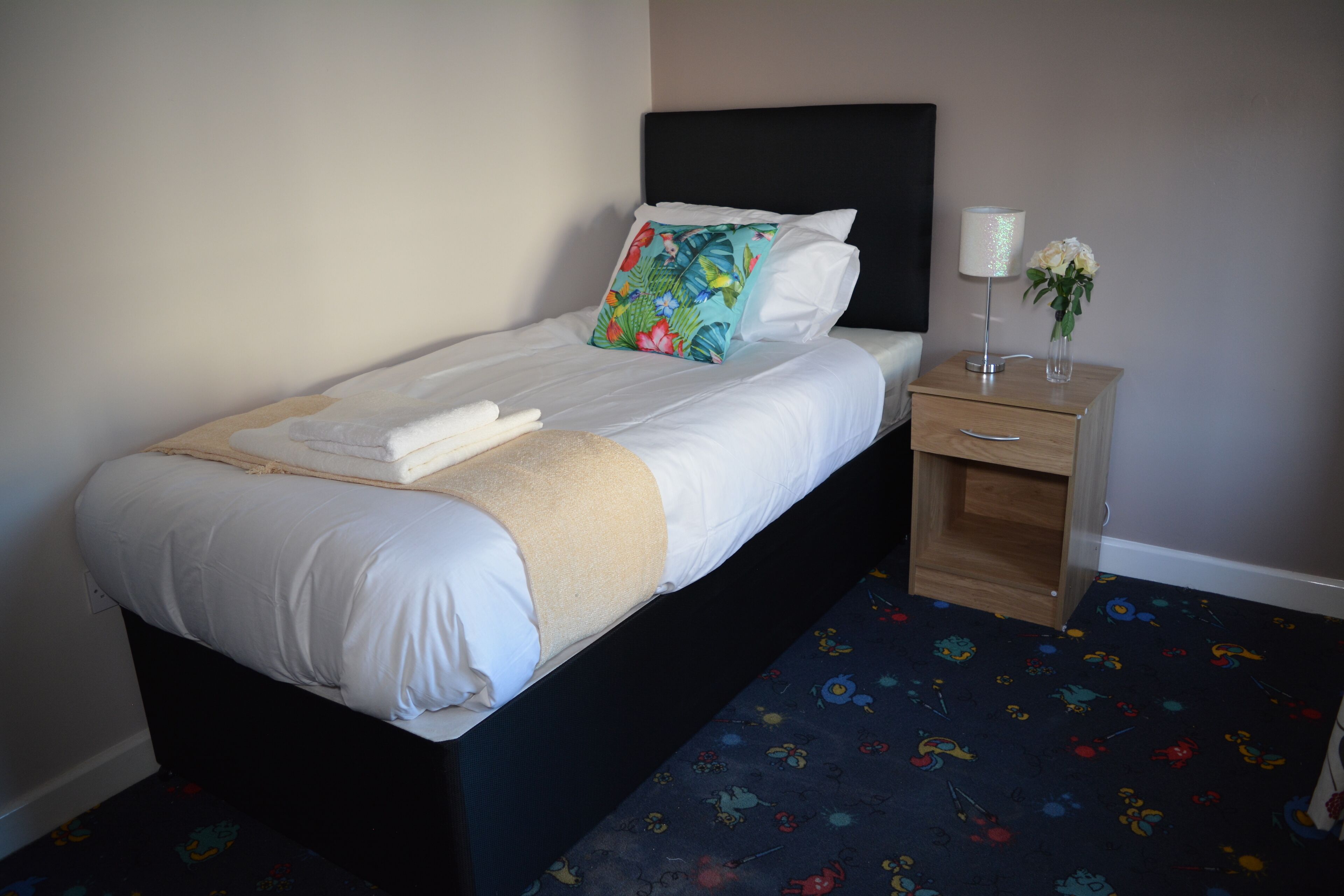 A neatly made bed with white linens and a beige throw, a nightstand with a lamp and a vase of flowers, and a carpet with a colorful pattern.