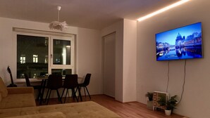A living room with a sofa, a dining table with chairs, a TV mounted on the wall, and a window with a view of buildings. - Central luxury apartment close to Prater / Messe / WU (Leopoldstadt)