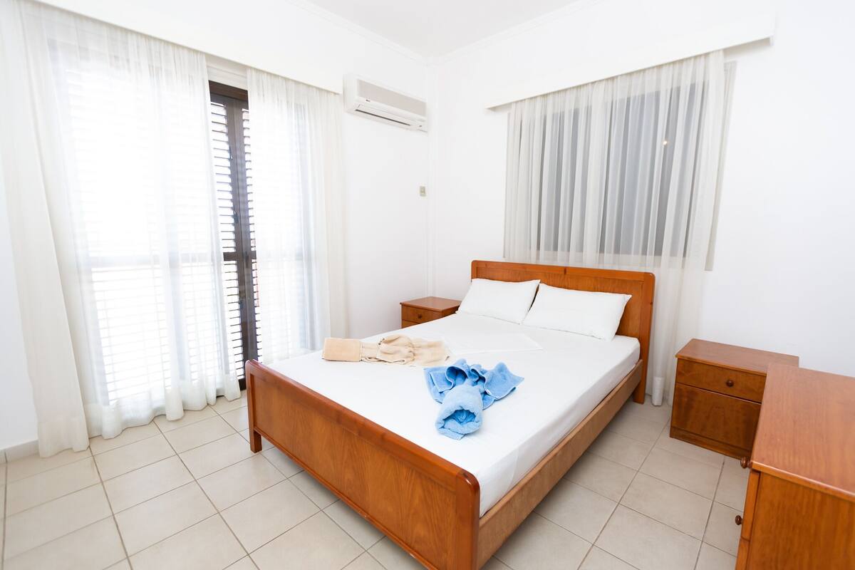 A clean, well-lit bedroom with a wooden bed, white linens, and a blue towel on the bed. There is a wooden nightstand and a small drawer unit beside the bed.
