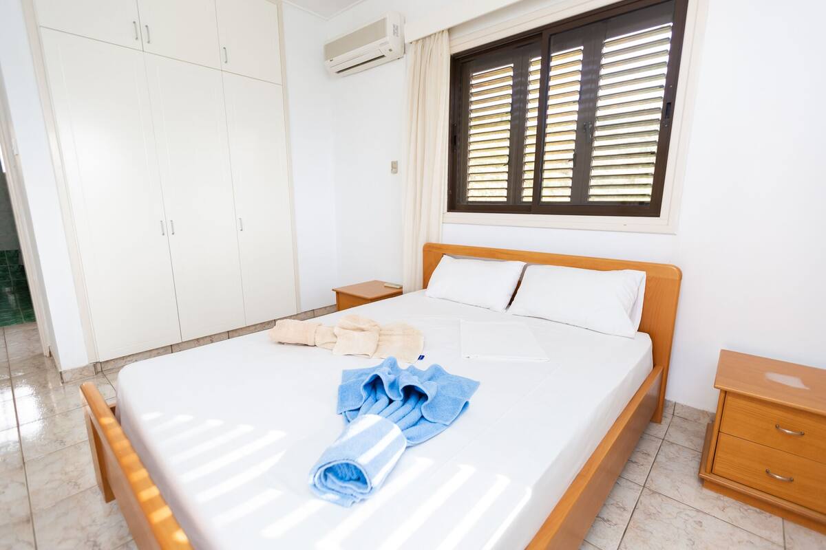 A hotel room with a bed, a wooden nightstand, a wardrobe, and a window with blinds.