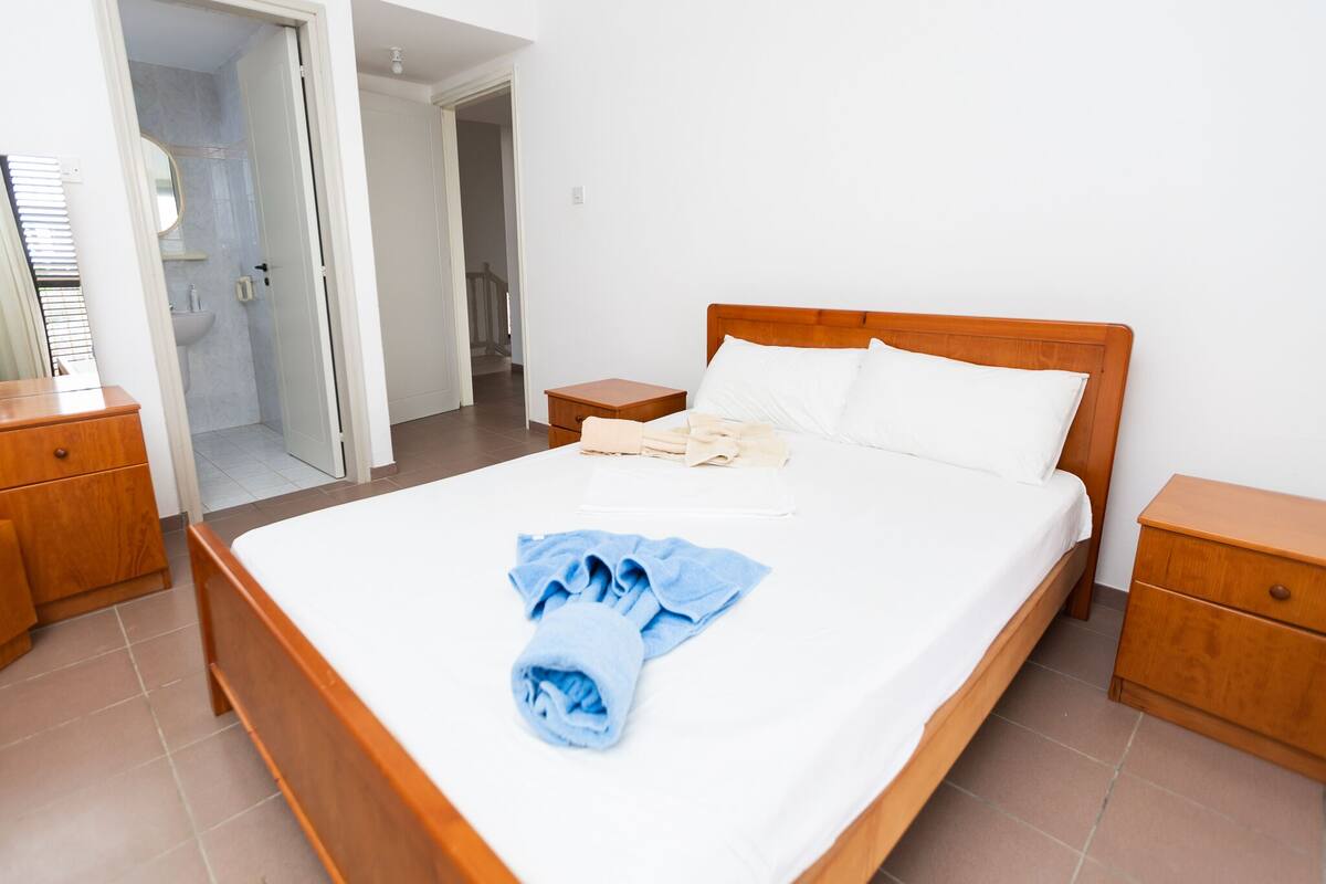 A neatly made bed with a blue towel and a white pillow, wooden nightstands, and a bathroom visible through an open door.