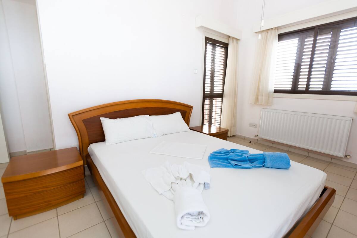 A neatly made bed with white linens and blue towels, a wooden nightstand, and a window with blinds.