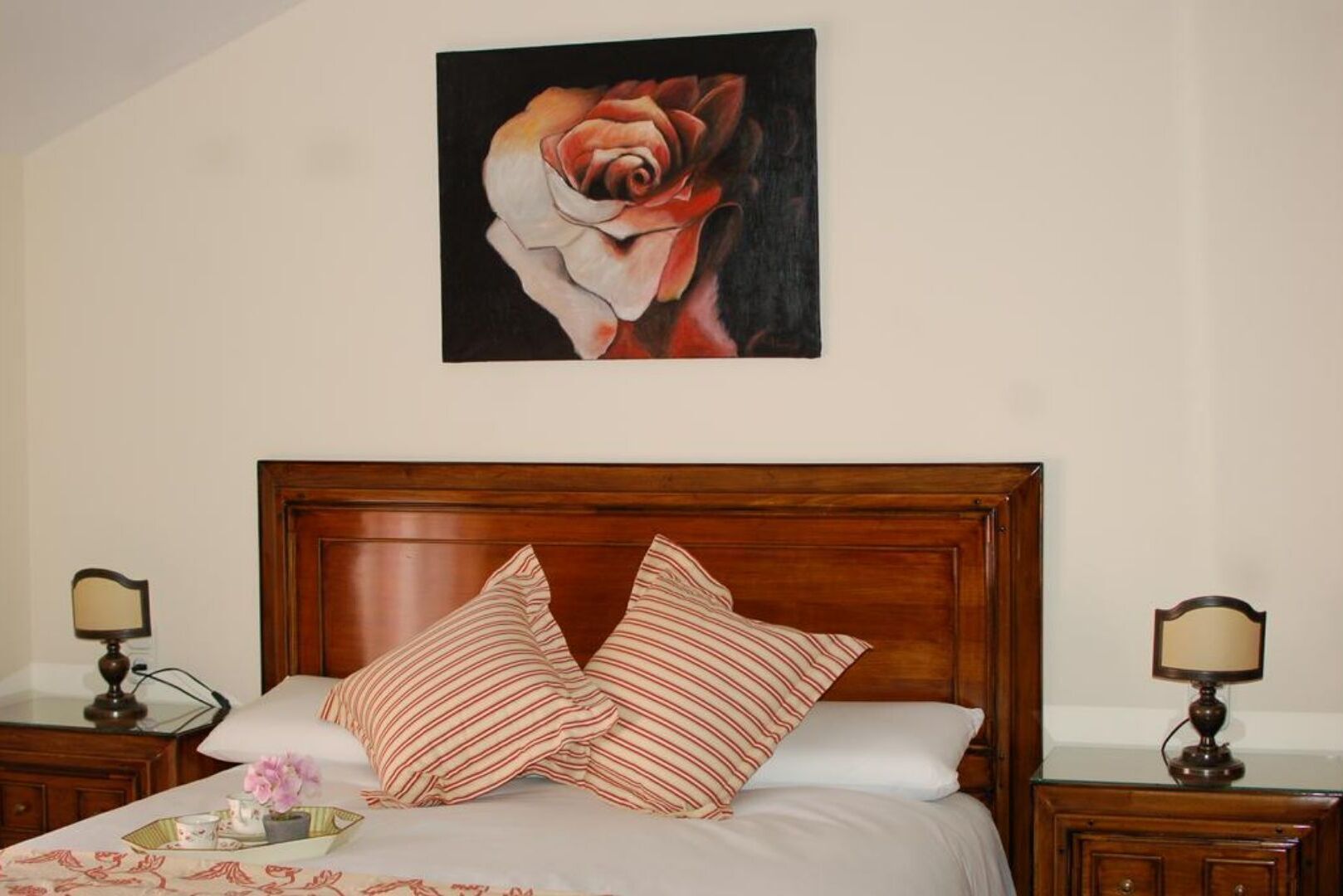 A bedroom with a wooden headboard, two bedside tables with lamps, a bed with striped pillows, and a painting of a rose on the wall.