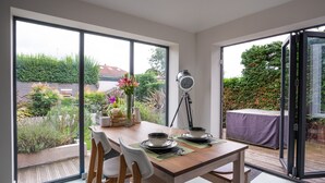 A dining area with a table set for two, large windows, and a view of a garden. - Contemporary Bright House 20 Min To Manc (Stockport)