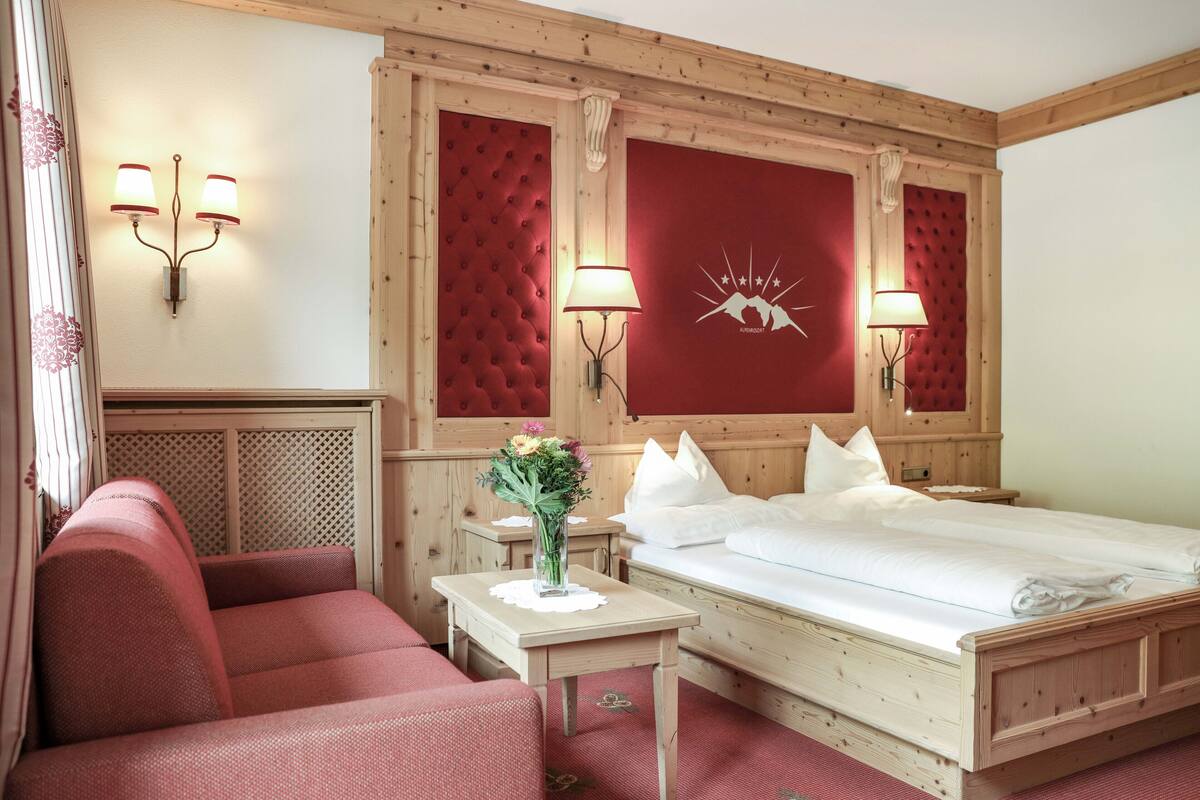 A hotel room with a wooden bed, a red upholstered sofa, a small table with a vase of flowers, and two wall-mounted lamps.