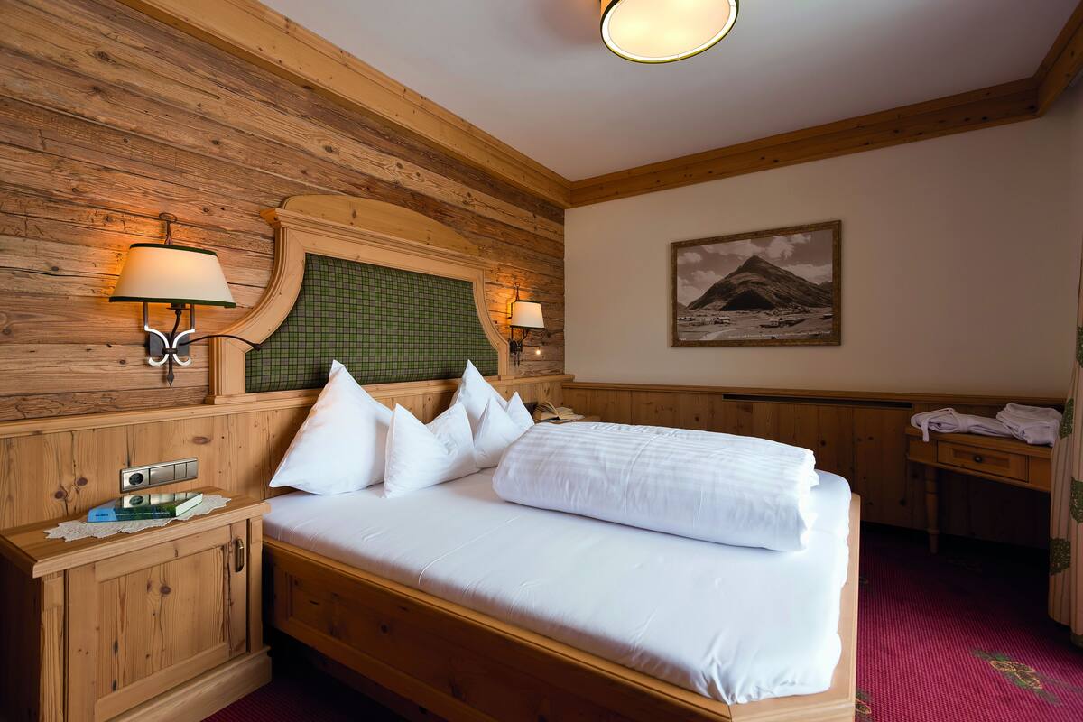 A wooden room with a bed, two wall-mounted lamps, a framed picture, and a wooden desk with towels.