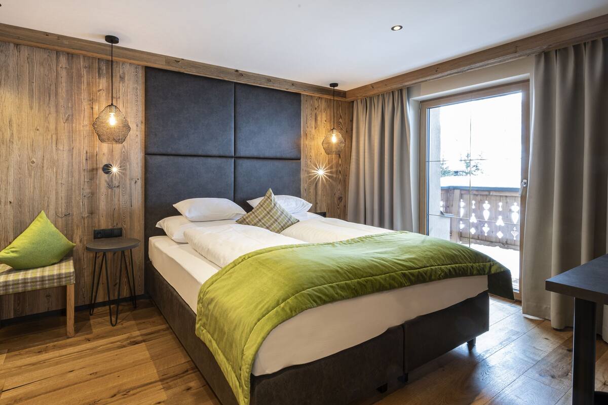 A hotel room with a wooden wall, a bed with white bedding, a green throw, a small table, and a chair.