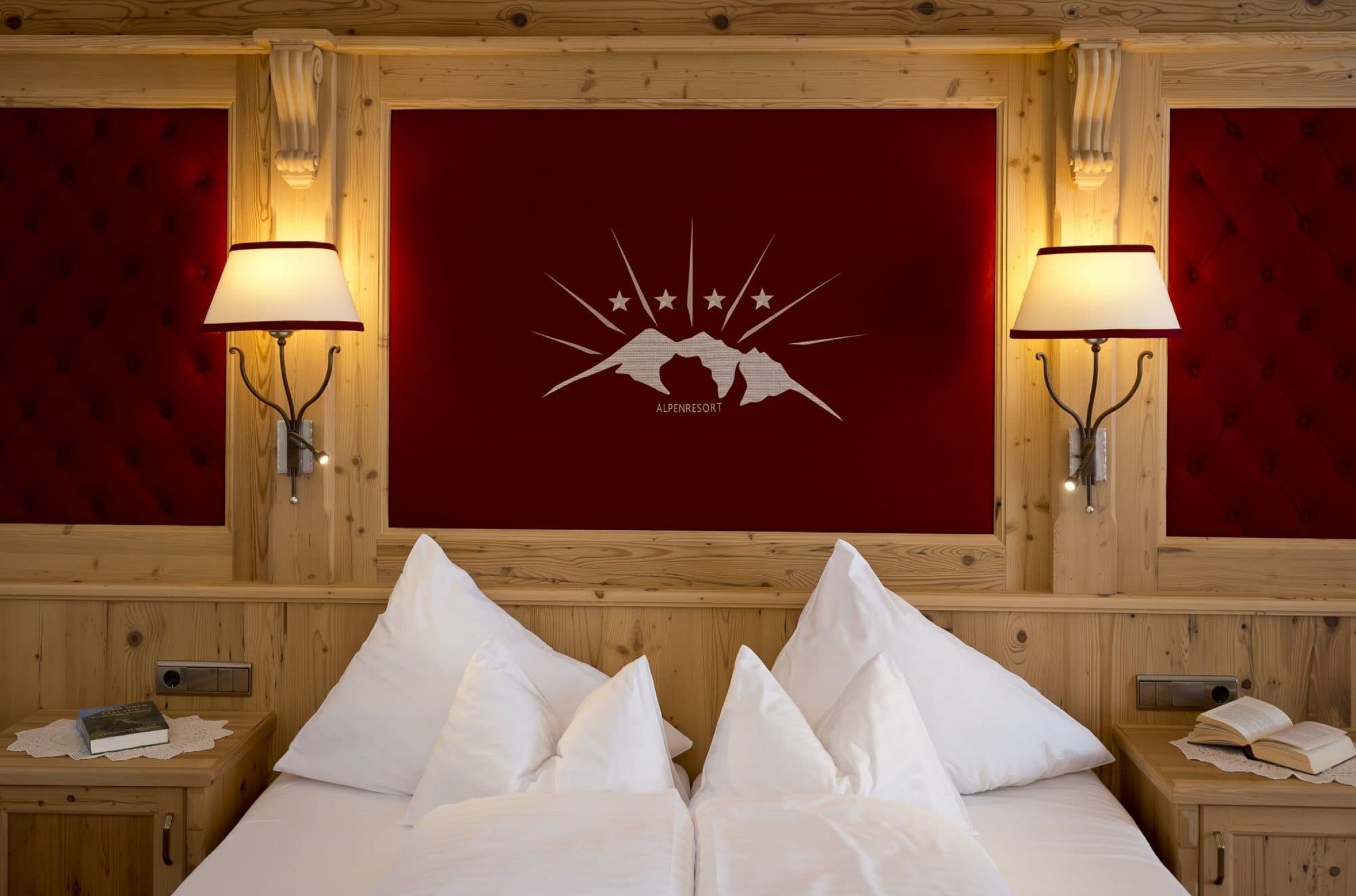 A hotel room with a wooden headboard, two bedside lamps, a bed with white linens, and a red wall with a mountain logo.