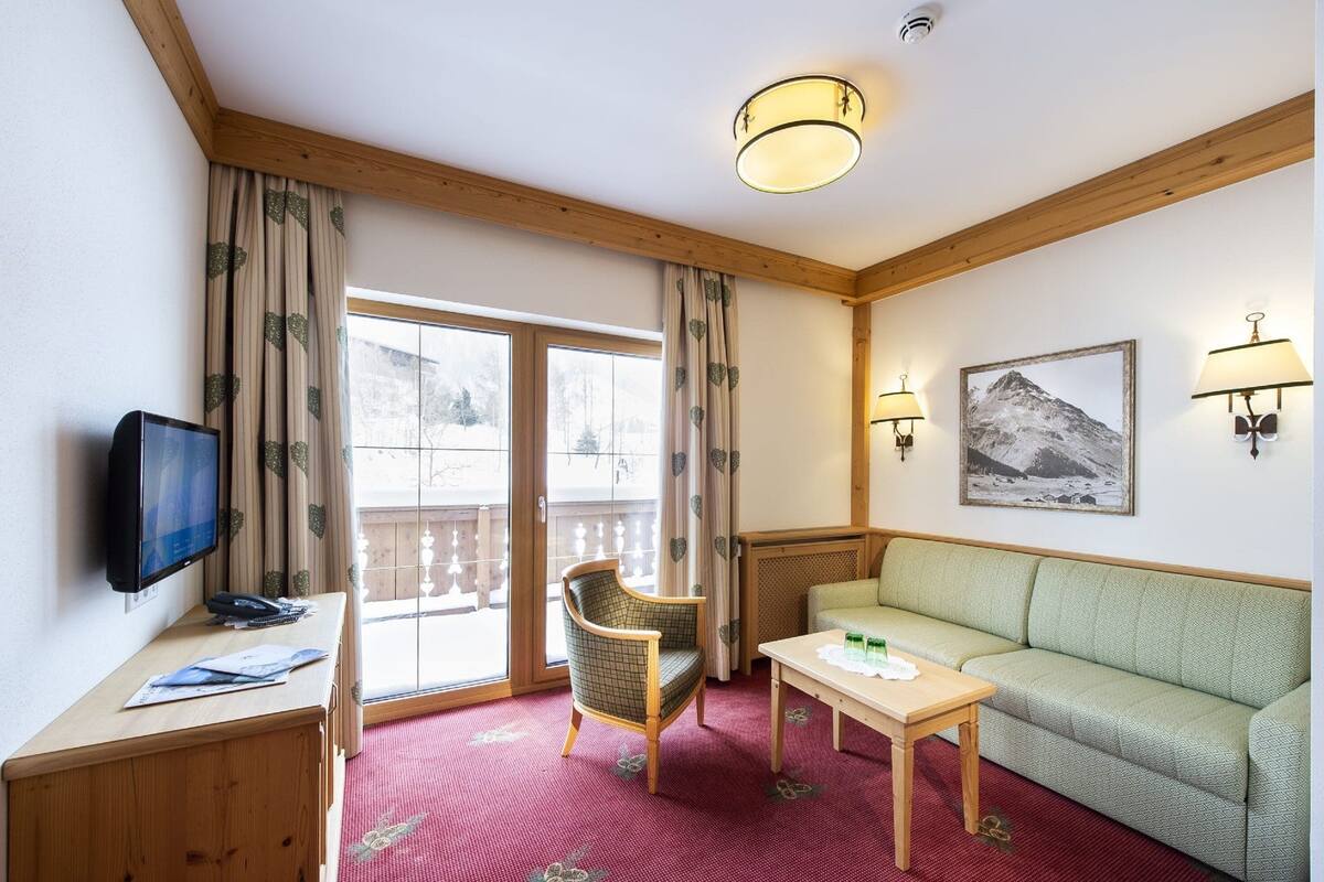 A hotel room with a wooden ceiling, a television, a desk, a chair, a sofa, a table, and a window with curtains.
