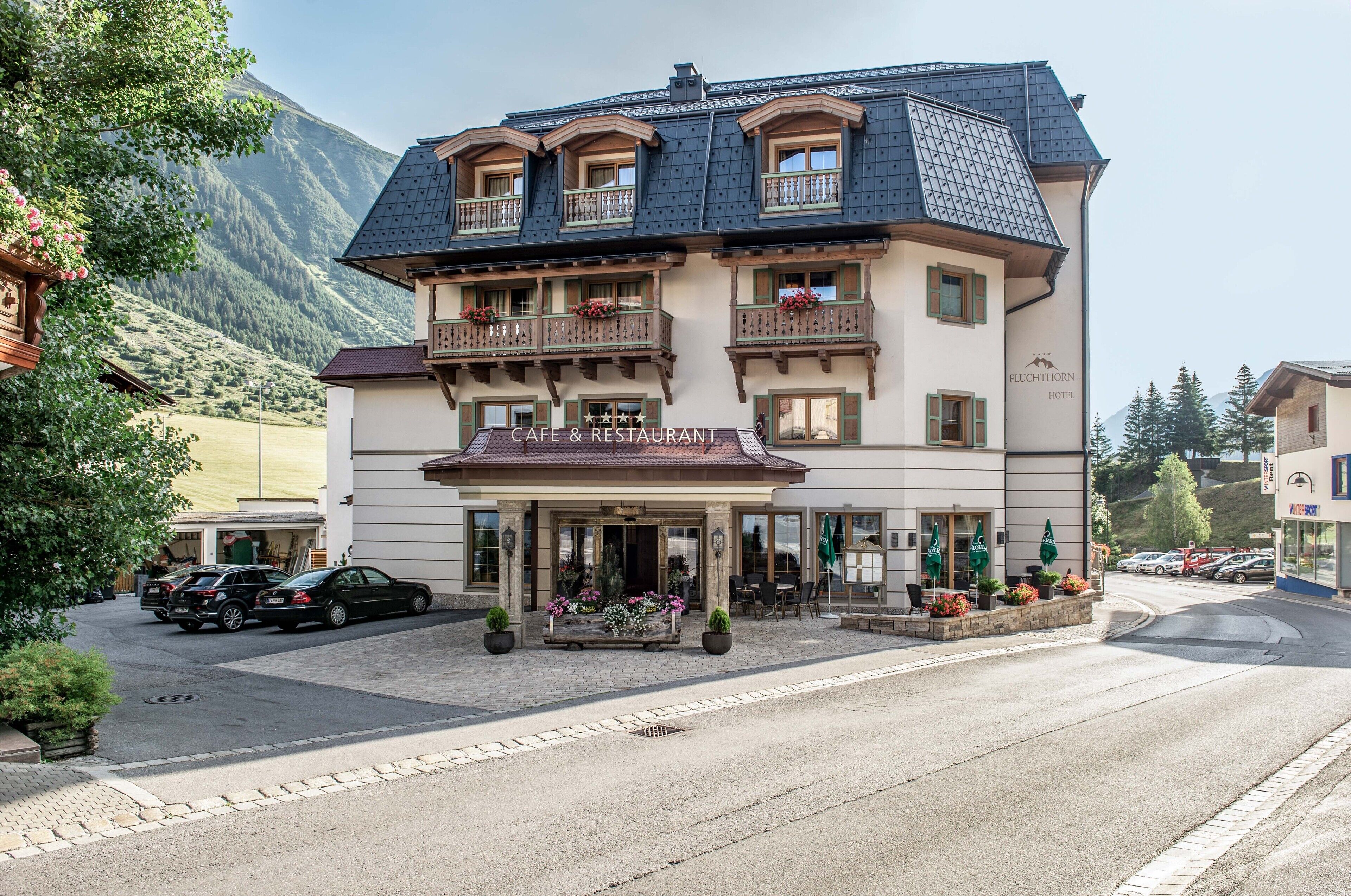Un hotel de varios pisos con estilo arquitectónico europeo tradicional, con balcones adornados con macetas de flores, un café y un restaurante en la planta baja, y un estacionamiento al frente.